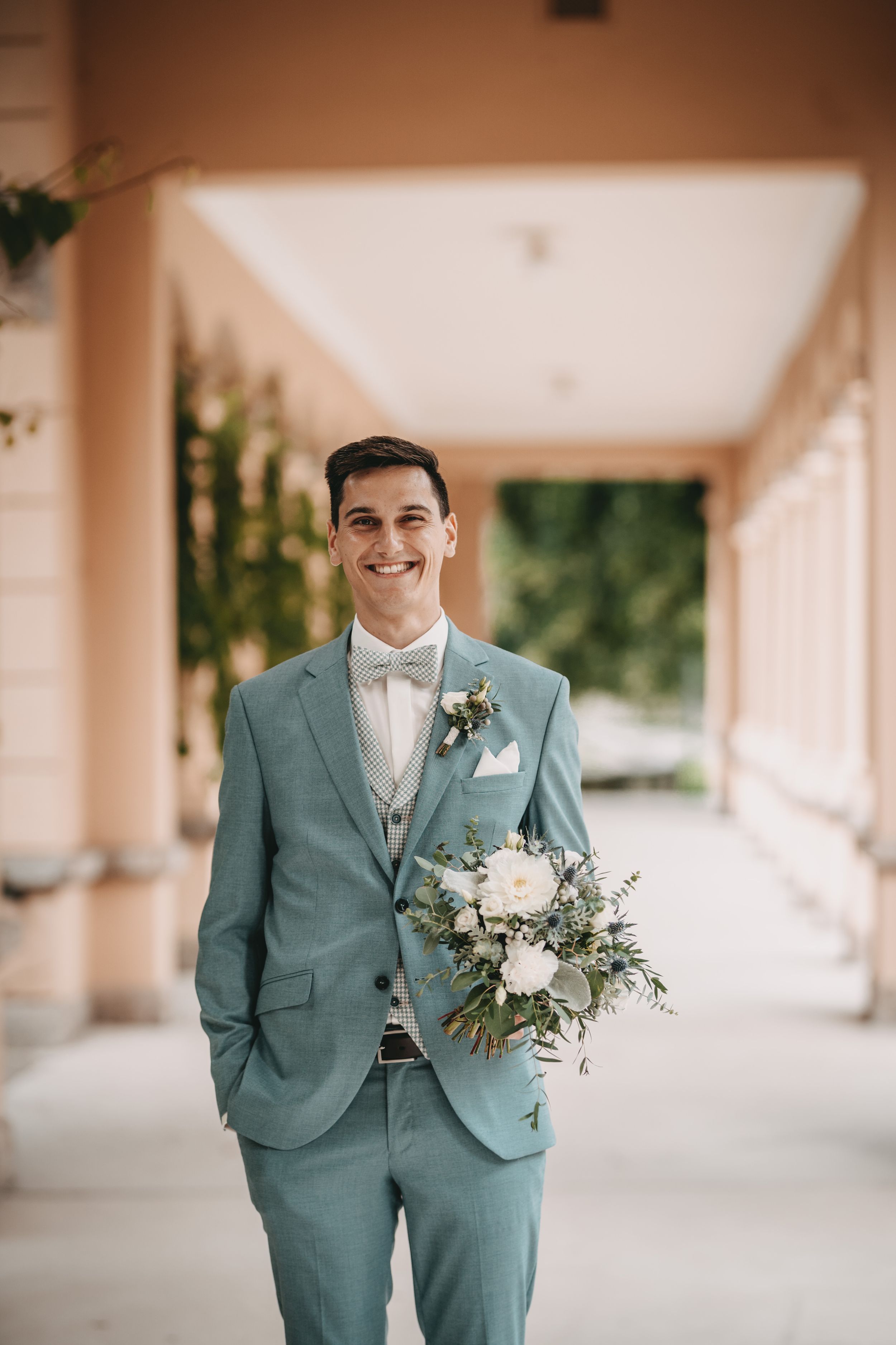 Groomsmen in sage green suits stand in a covered outdoor walkway with white floral boutonnieres.