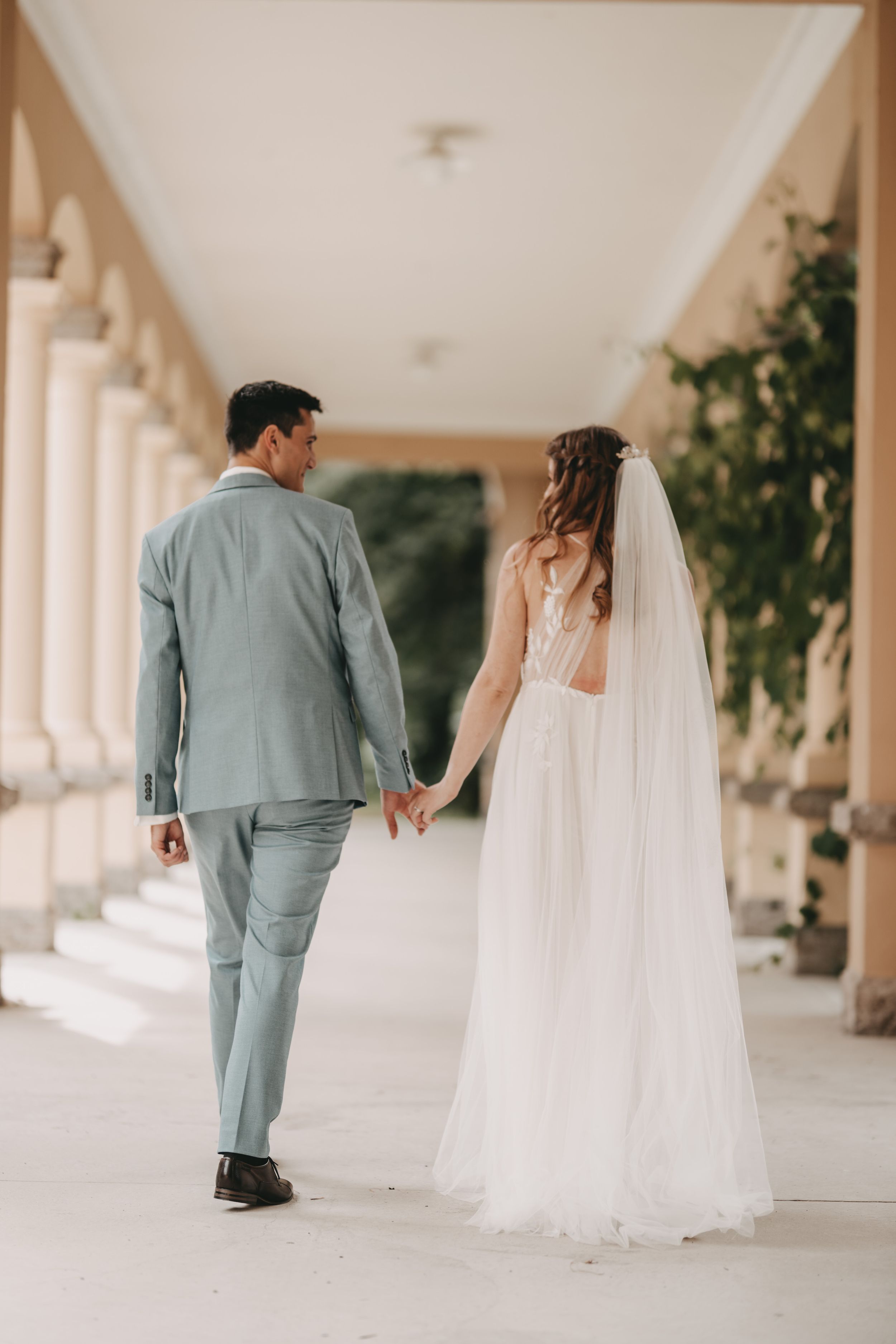Bride in flowing white gown follows groom down elegant colonnade.