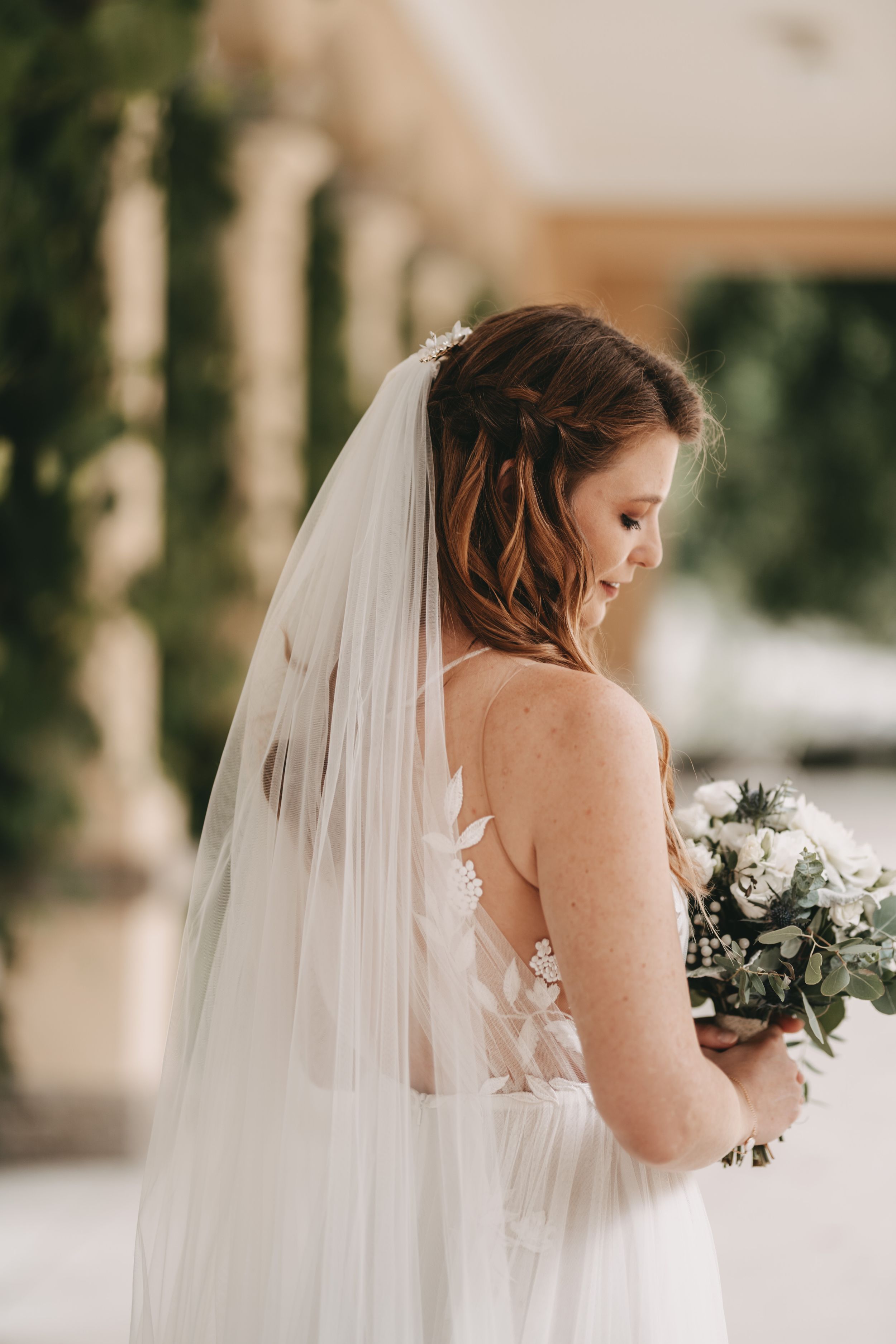 Bride in white lace gown holds delicate bouquet while posing in elegant colonnade.