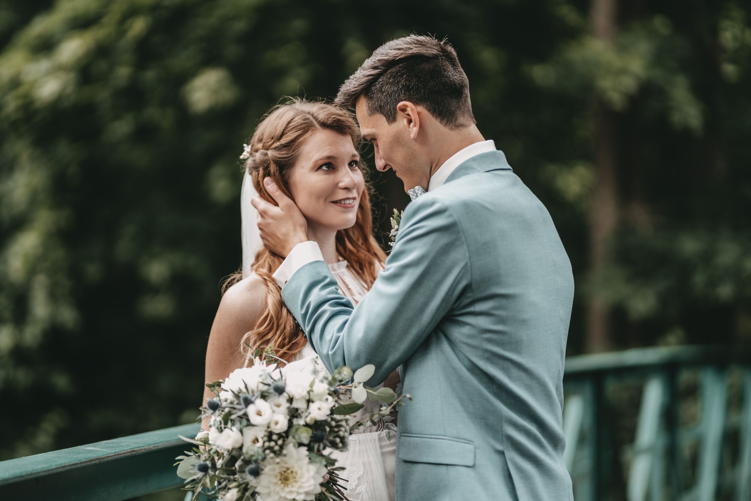 Wedding couple poses against lush greenery in light blue suit and white dress.