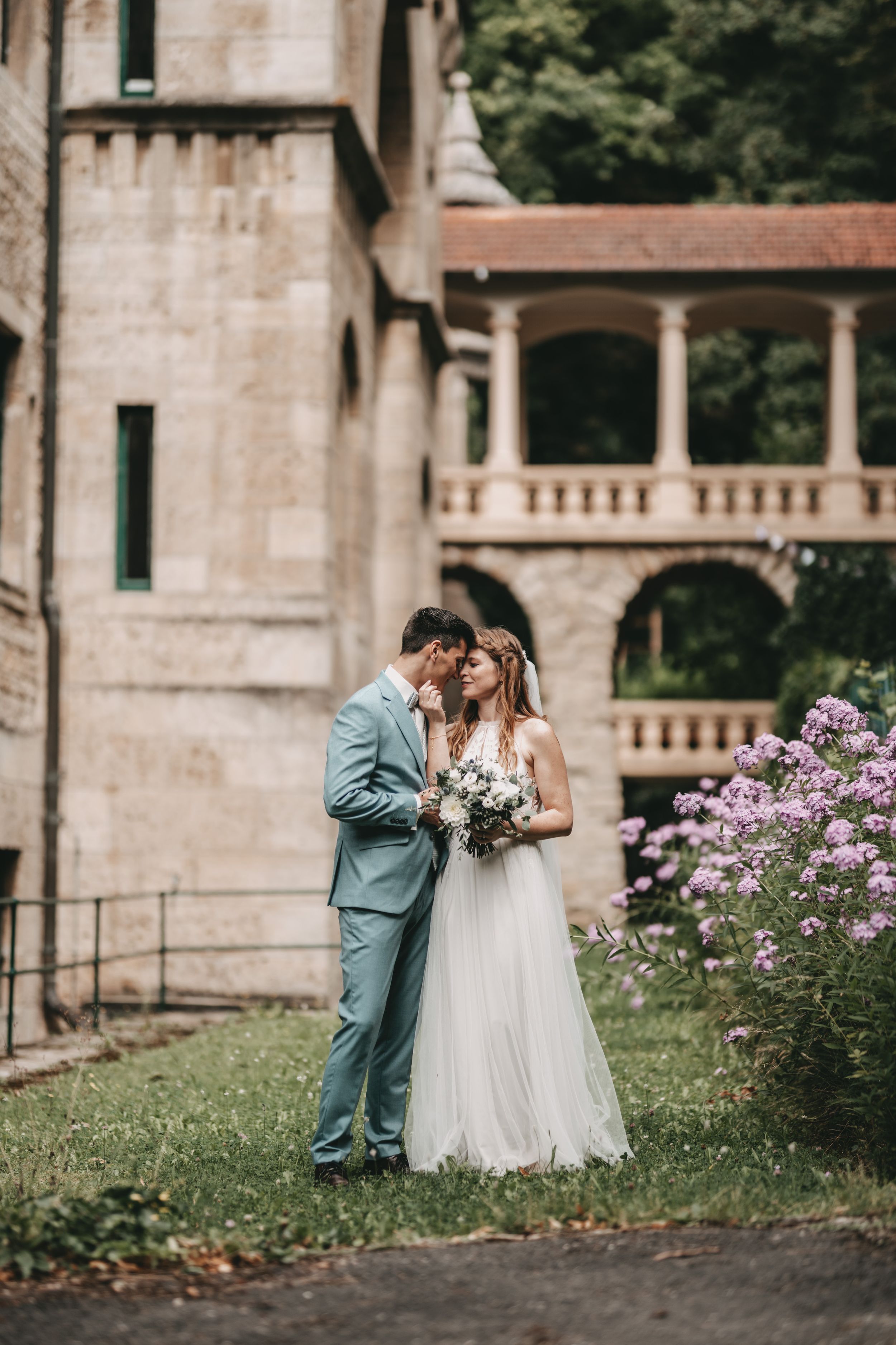 Wedding couple poses near stone building with purple flowers blooming in garden.