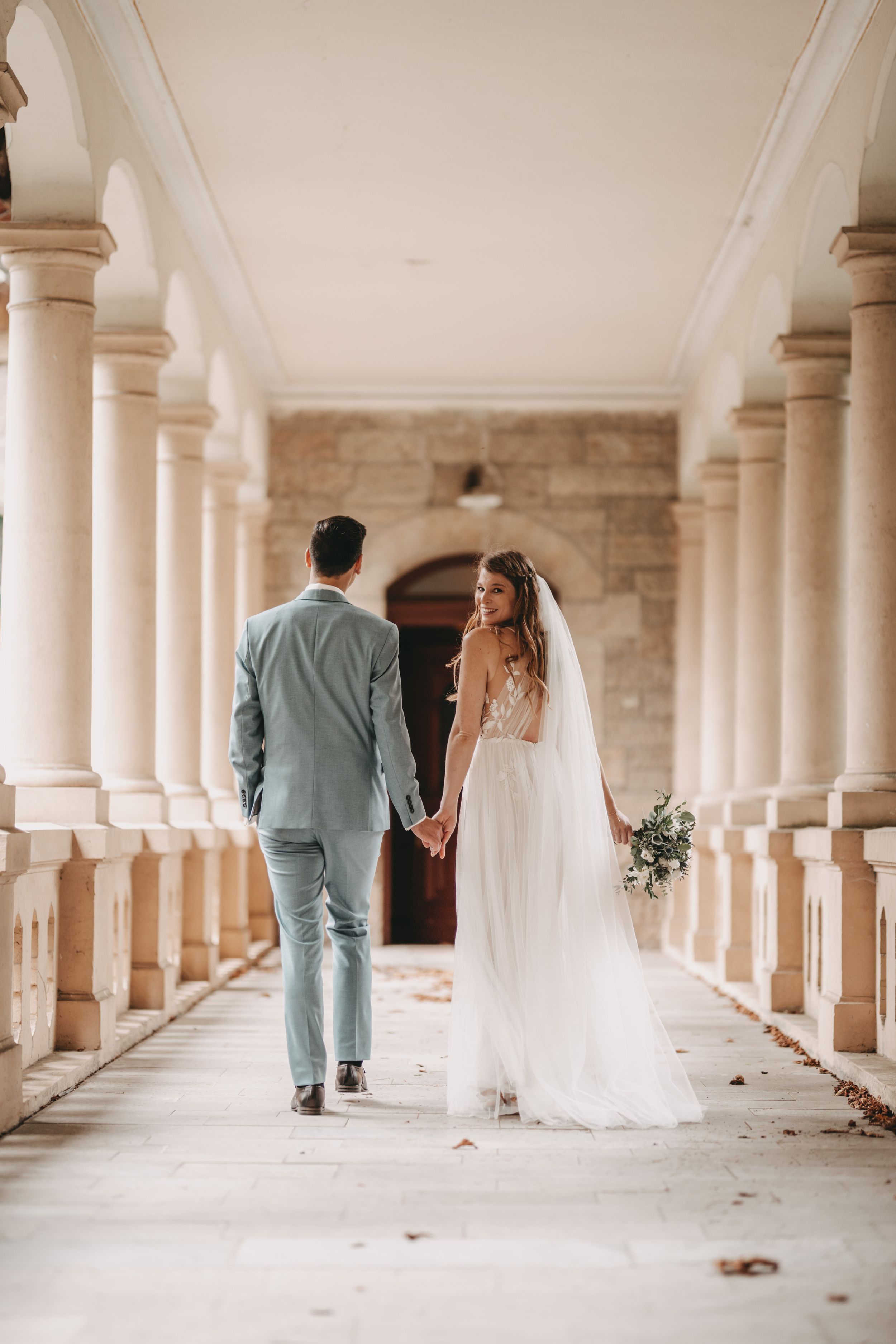 Elegant wedding couple walks through historic colonnade with white pillars.