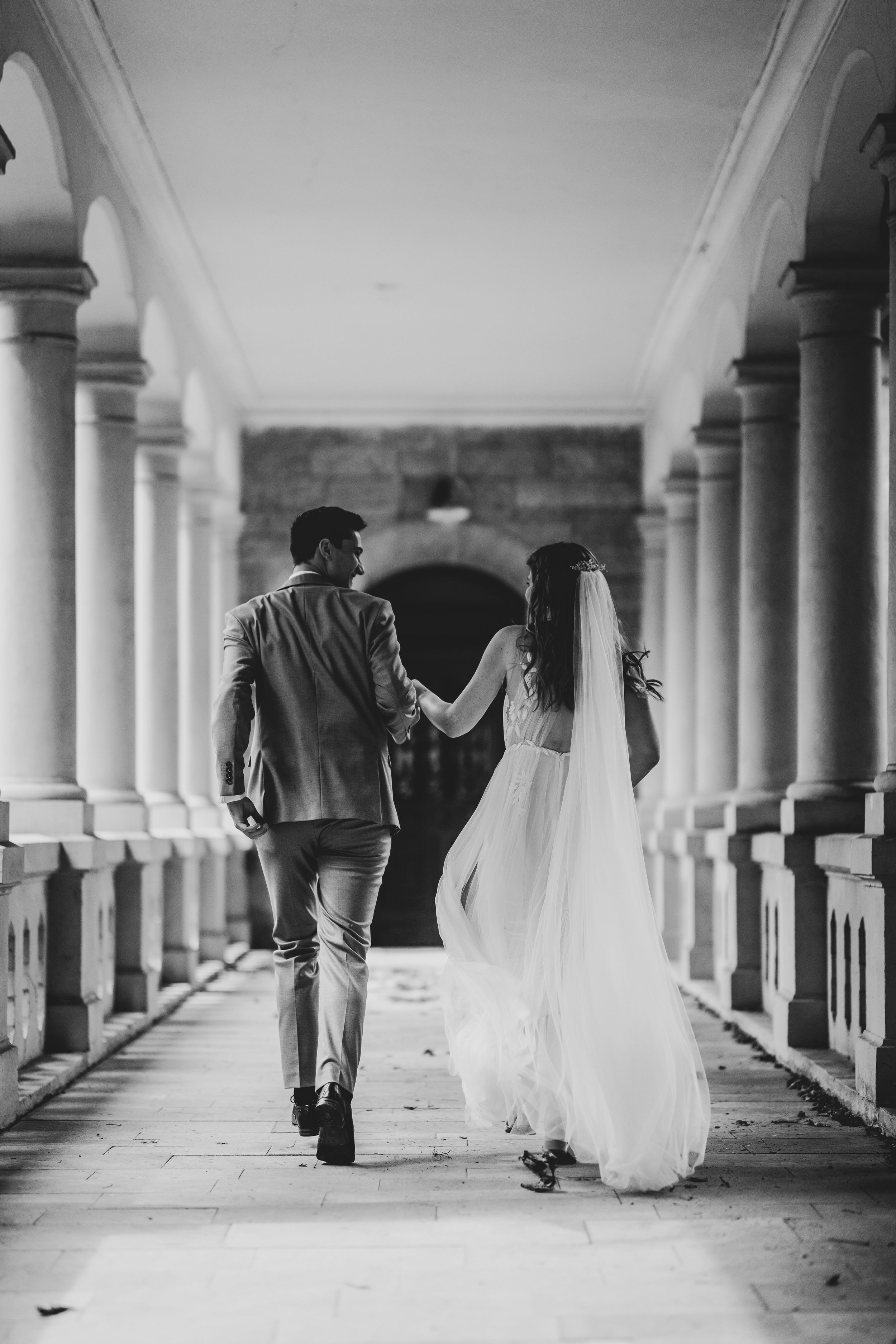 Black and white wedding photo of couple walking through classic columned hallway.