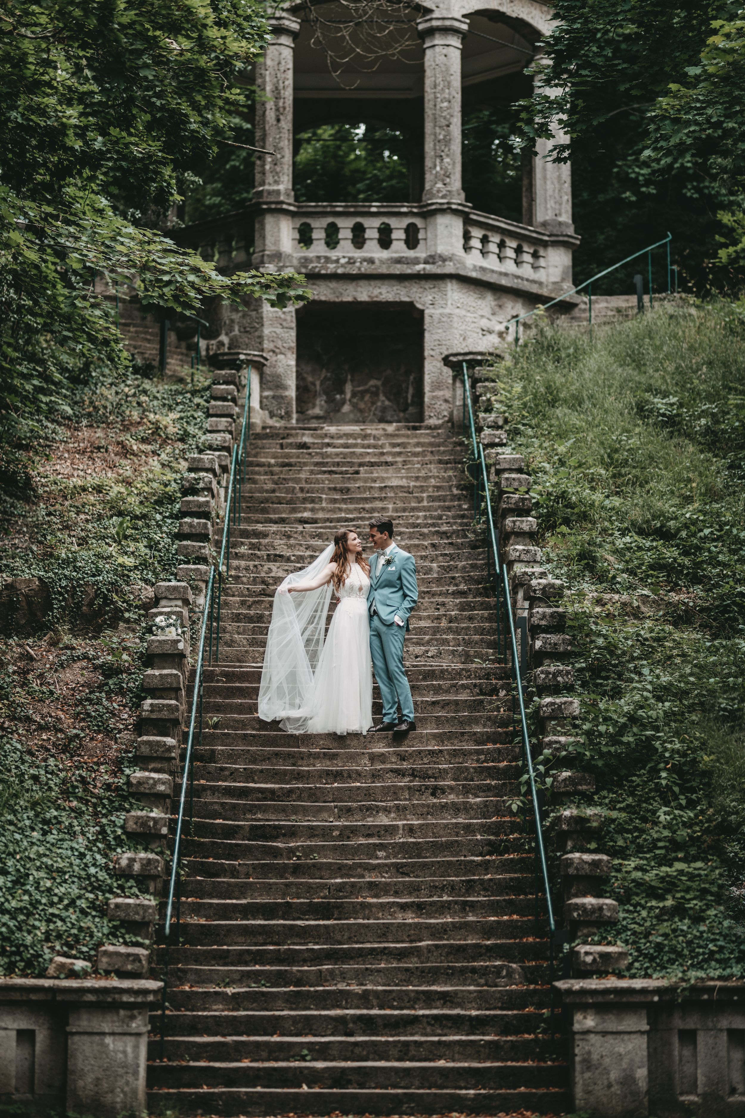 A couple in wedding attire embracing on grand stone steps leading to an ornate gazebo.