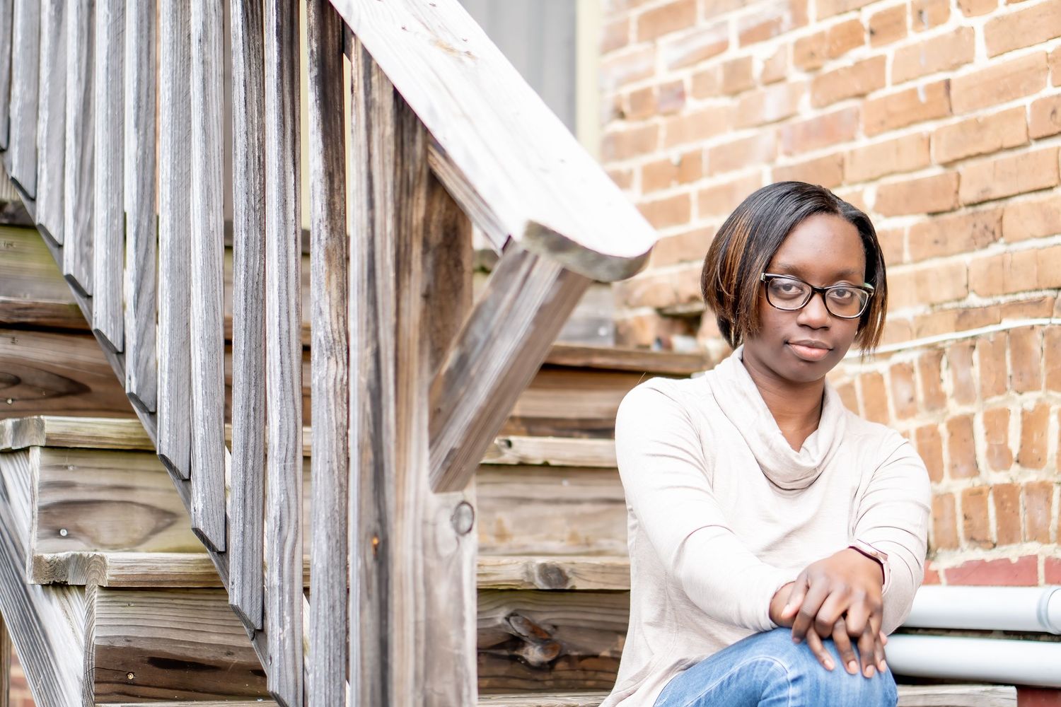 Person in glasses and white sweater sits thoughtfully on wooden stairs against a brick wall backdrop.