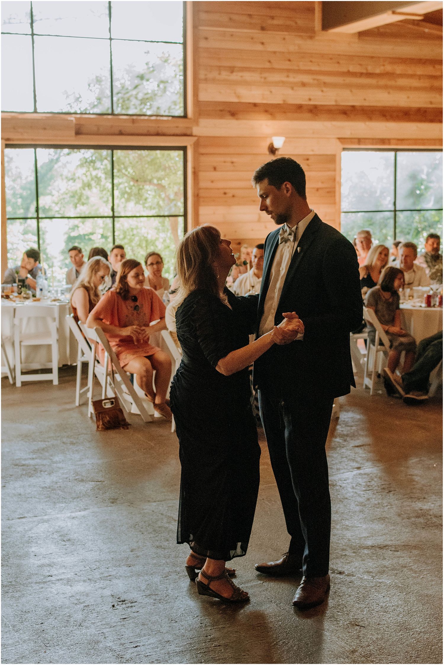 A couple sharing a dance in a rustic venue with large windows and white chairs.