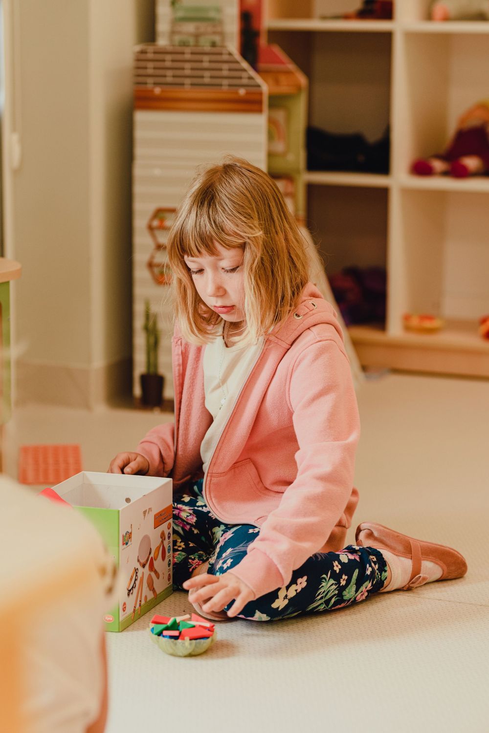Child in pink sweater lying on floor playing with toys.