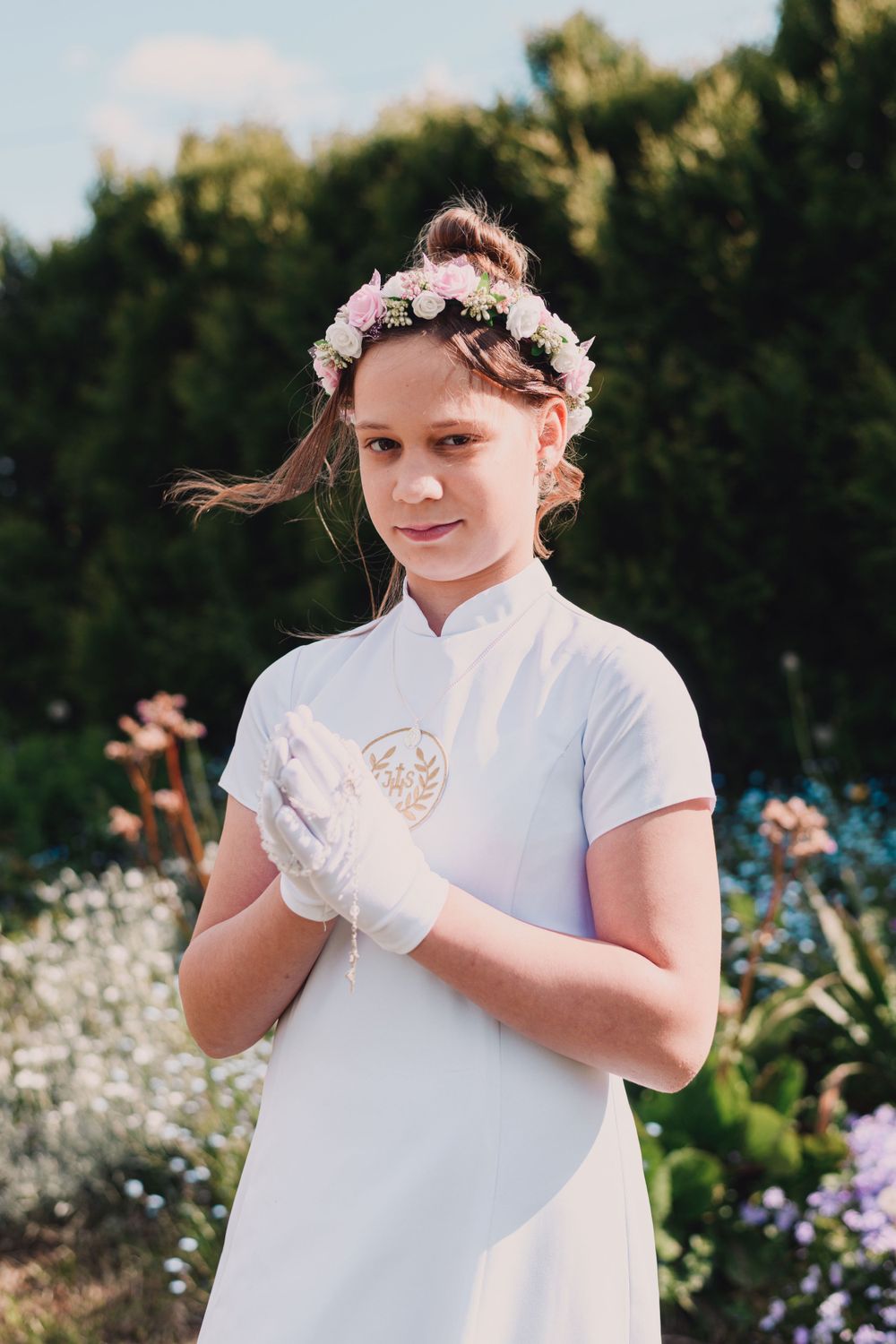 Young child wearing white dress and floral crown poses in garden with white gloves.