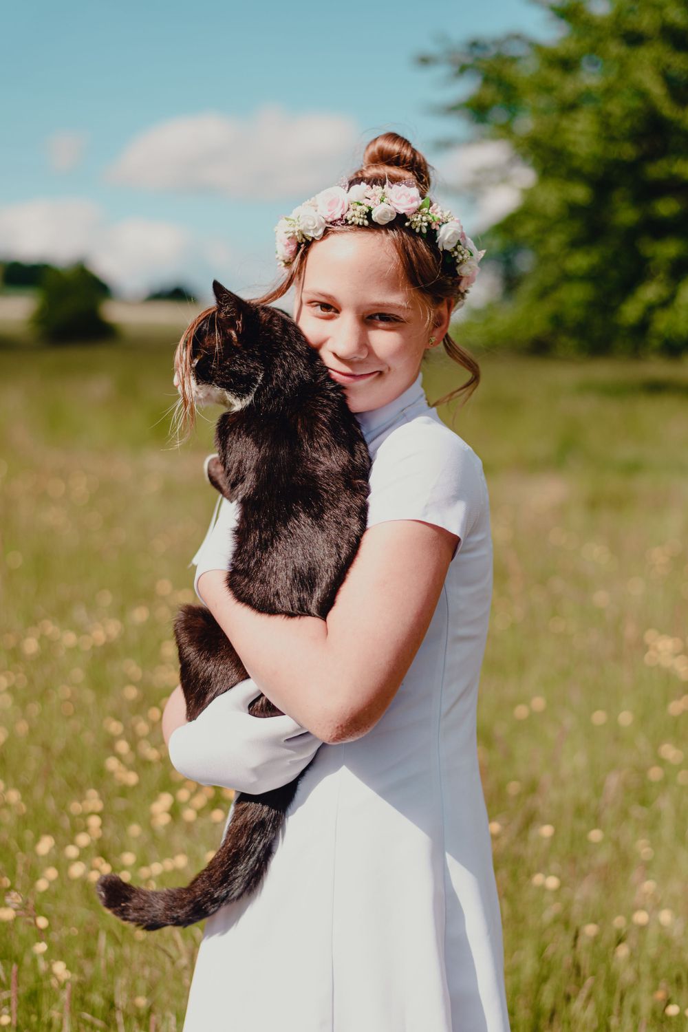 Child in white dress holding black cat in a blooming meadow.