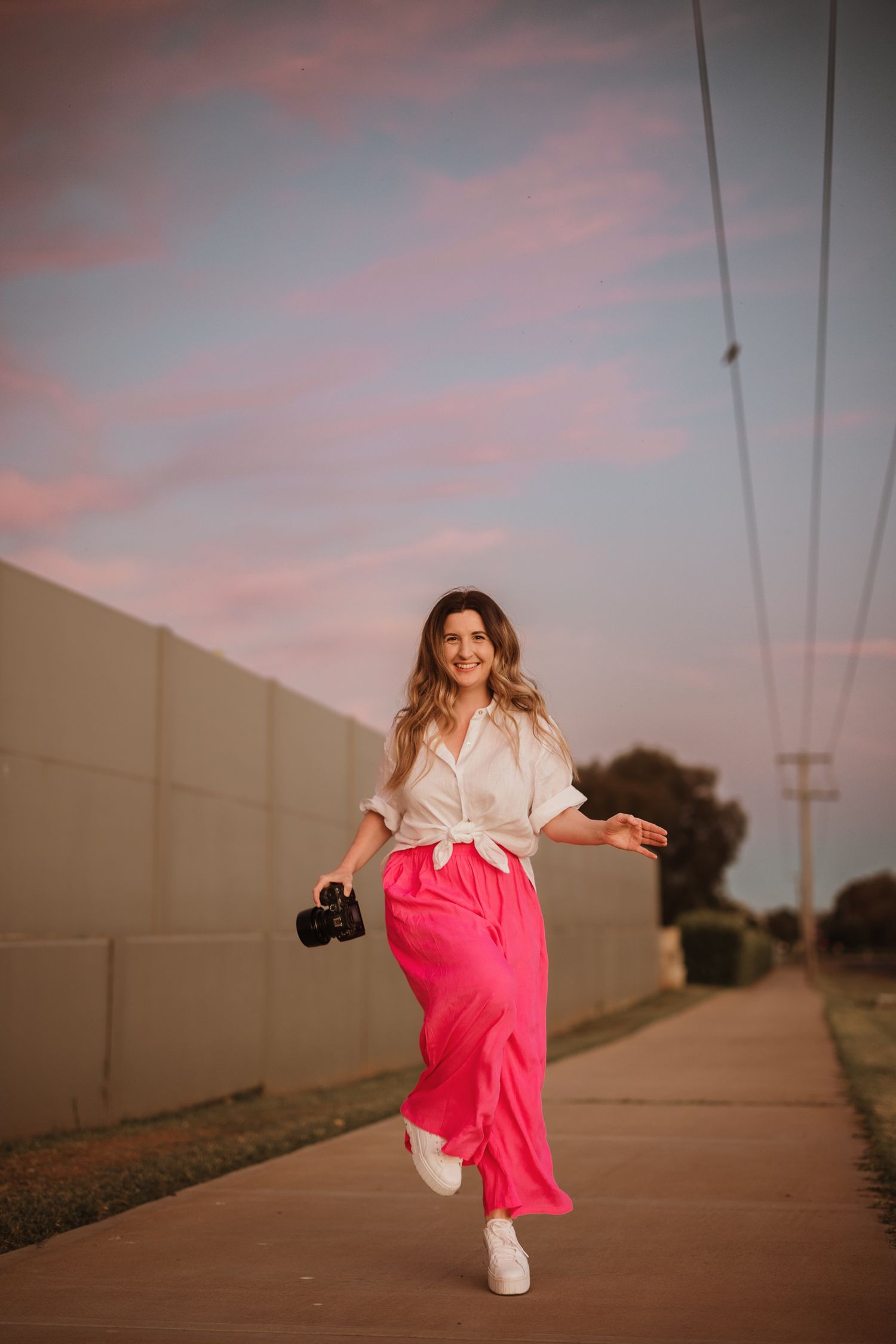 Photographer skipping down path in pink pants and white shirt with camera at dusk with pink sky