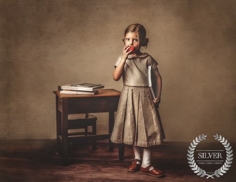 Young girl in vintage dress eating apple by school desk, silver award winning photo.