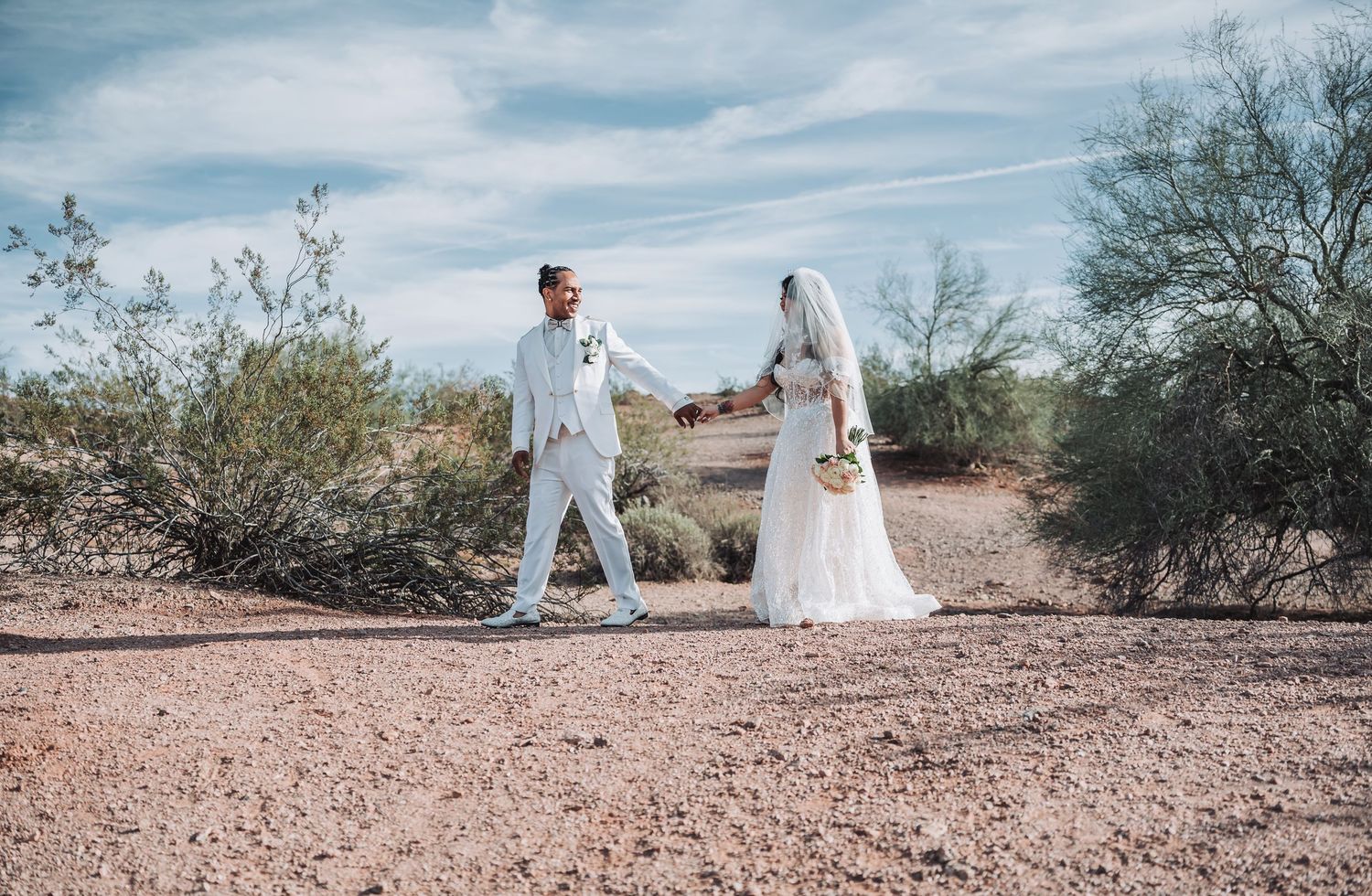 A wedding couple holds hands while walking on a desert path surrounded by desert brush.