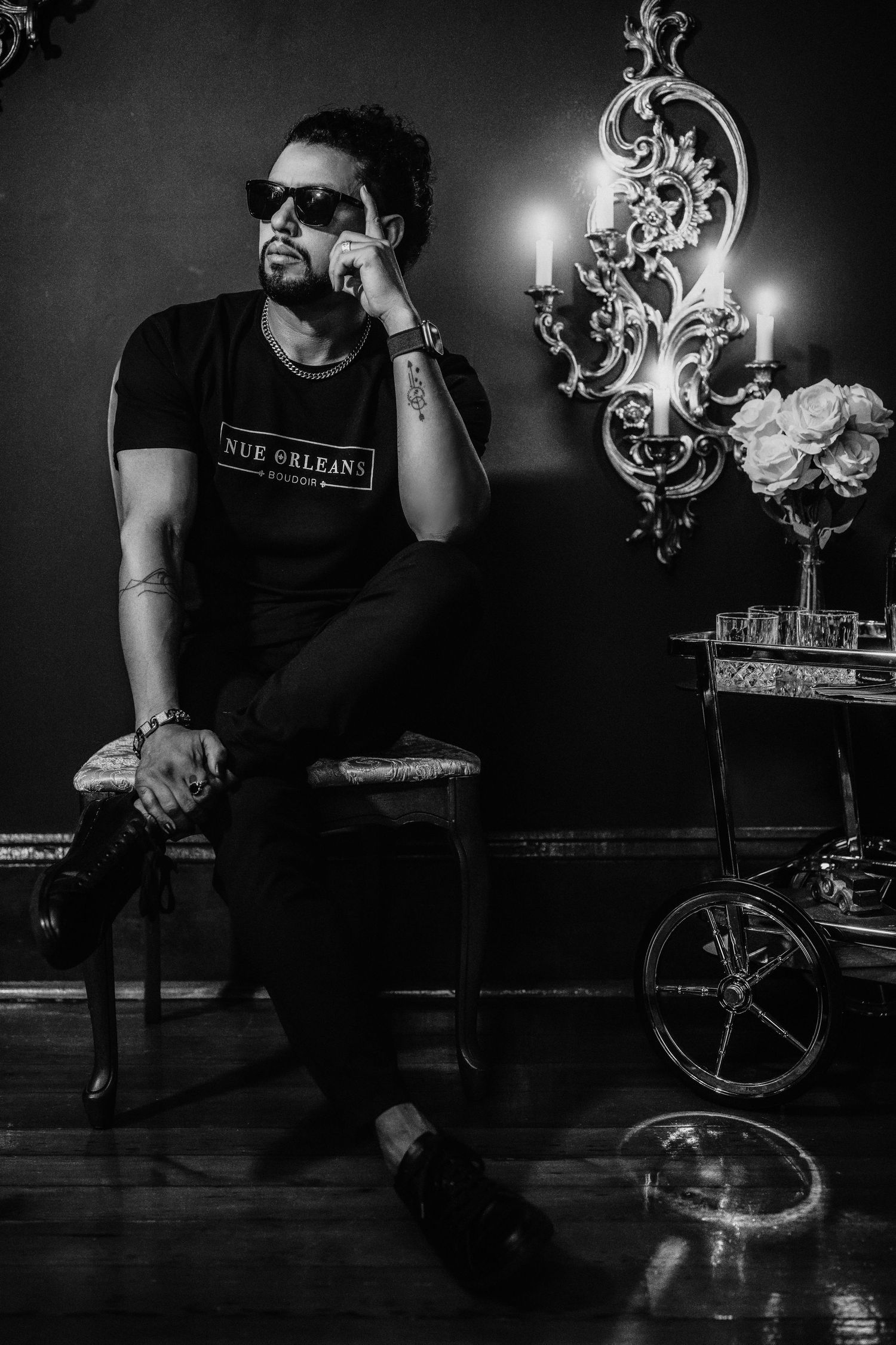 Moody portrait of someone in sunglasses seated on a chair next to ornate wall decor and candles.
