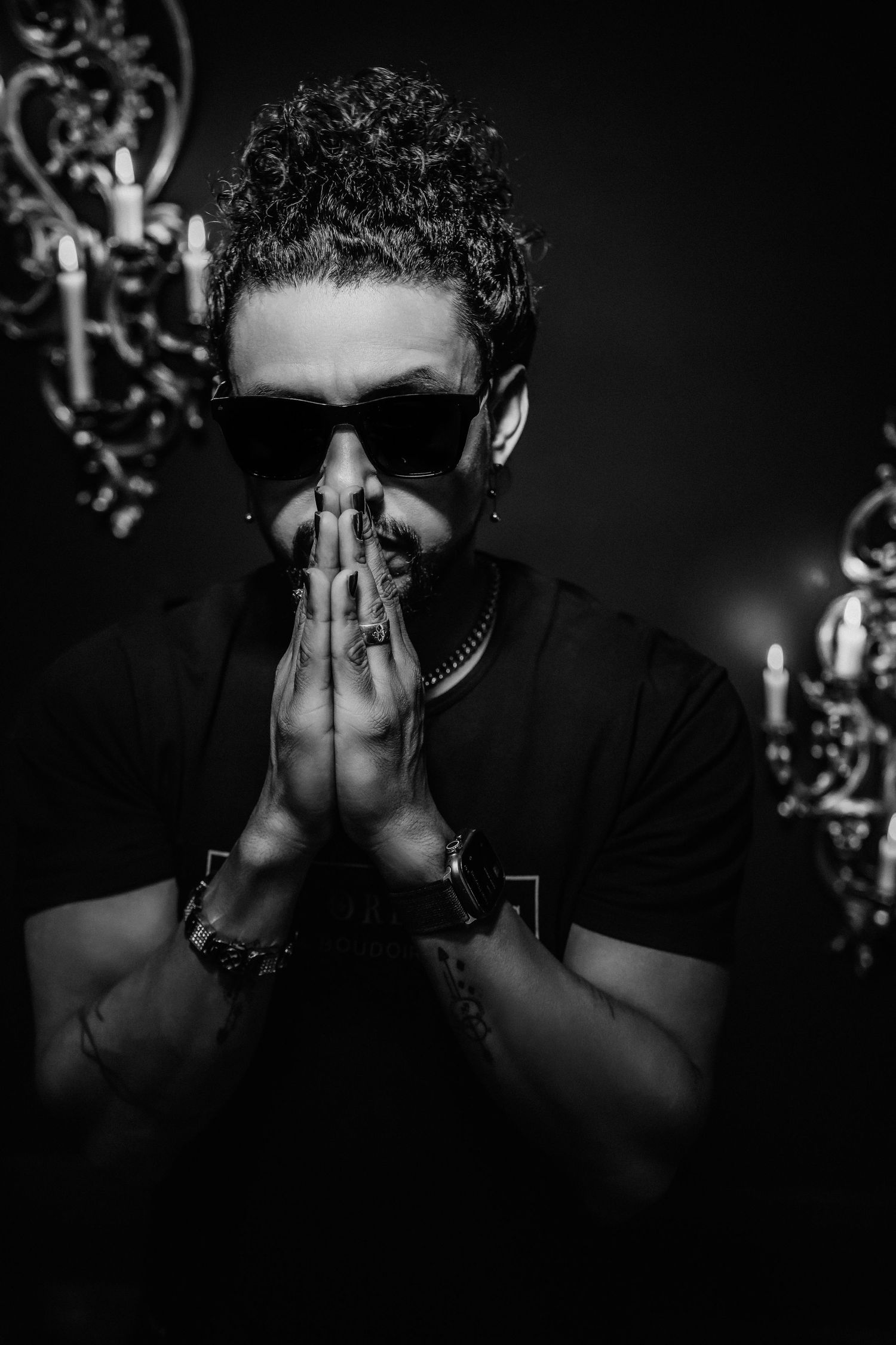 Dramatic black and white portrait of someone in sunglasses making a prayer gesture.