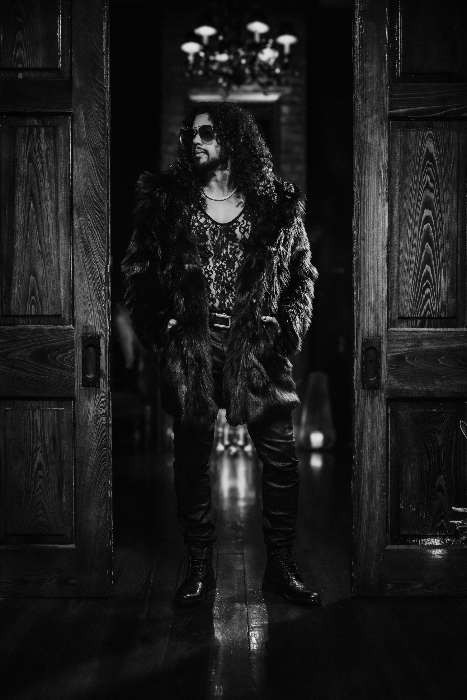 Atmospheric photo of someone in a fur coat standing between wooden doors with chandelier lighting.