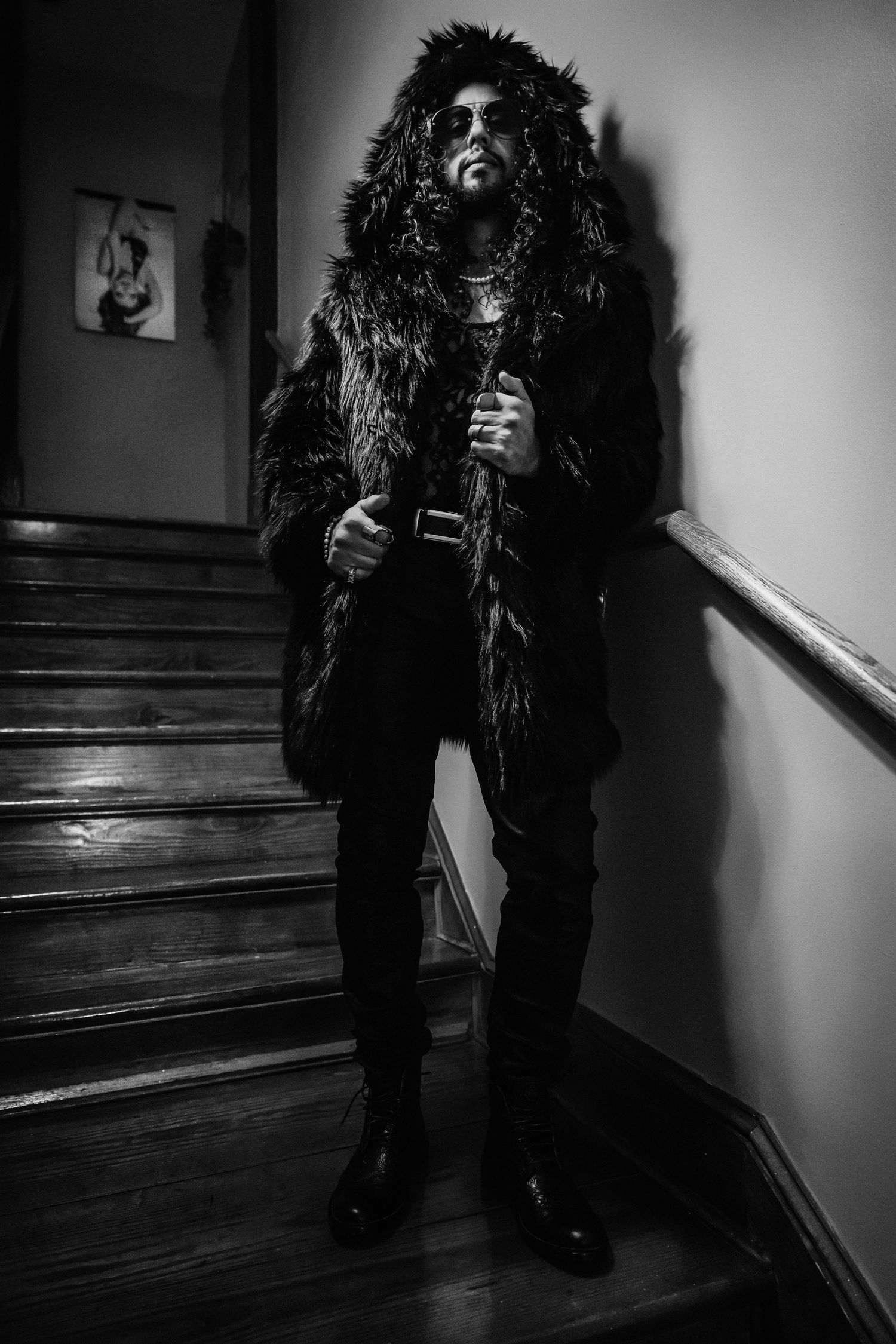 Dark atmospheric portrait of a person wearing a long furry coat posing on wooden stairs in black and white.
