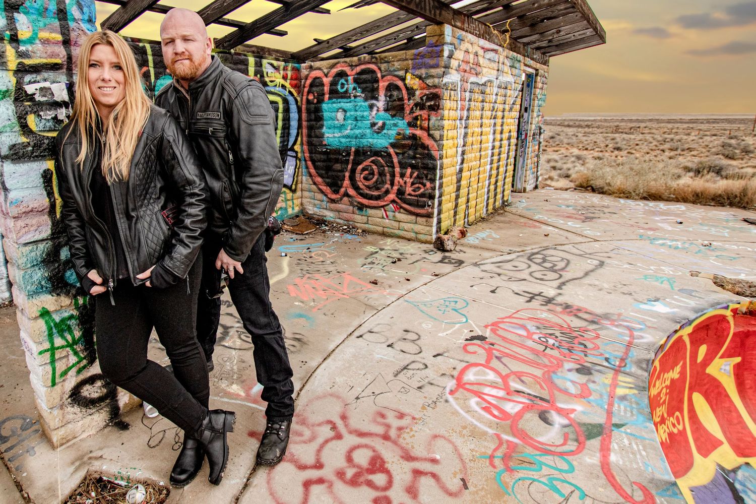 Two people in leather jackets stand next to a graffiti-covered structure in a desert landscape.