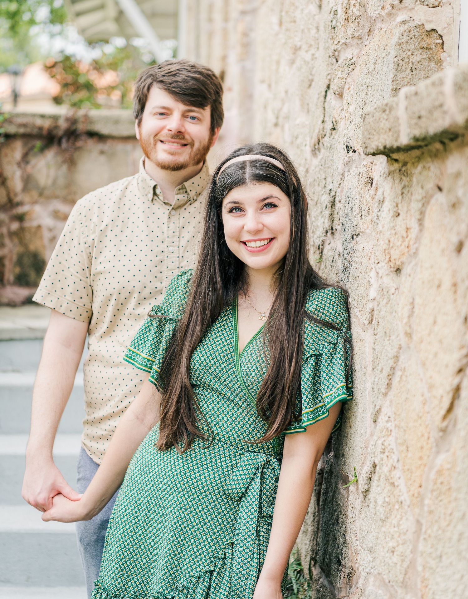 A couple poses against a stone wall, wearing coordinating outfits with green and beige patterns.