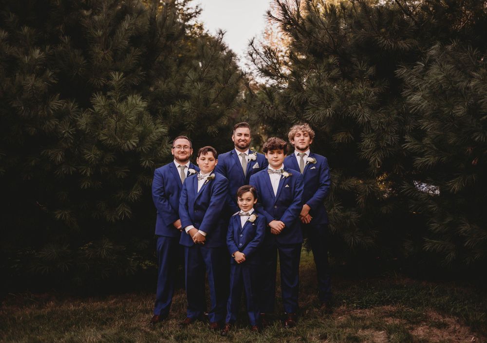 Groomsmen in matching navy suits pose together in front of pine trees.