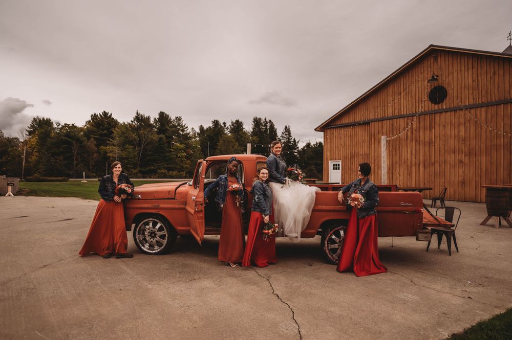 Wedding party gathered around vintage orange truck outside rustic barn.