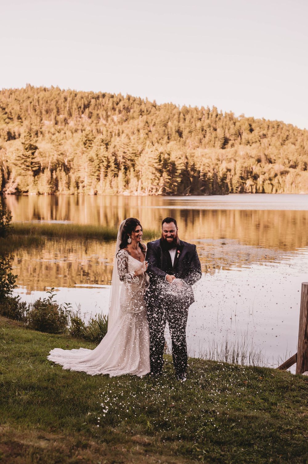 Wedding couple shares celebratory moment with confetti by serene lake at sunset.
