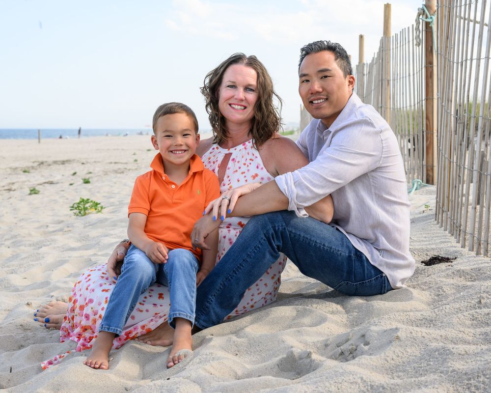 Family sitting together on sandy beach with wooden fence in background.