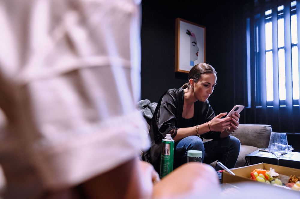 Person on couch checking phone with beverages and snacks on table in dark room with window light.