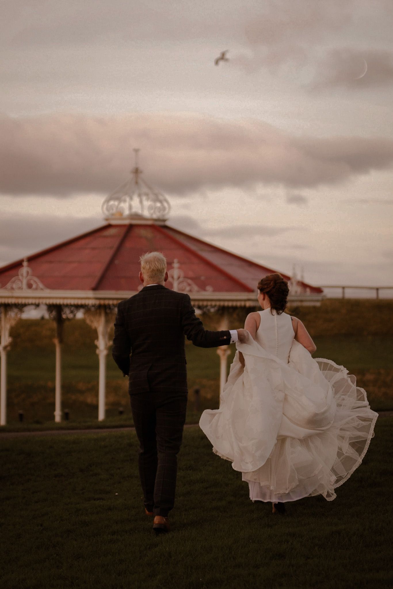 Couple walks towards red-roofed gazebo during romantic sunset wedding photoshoot.