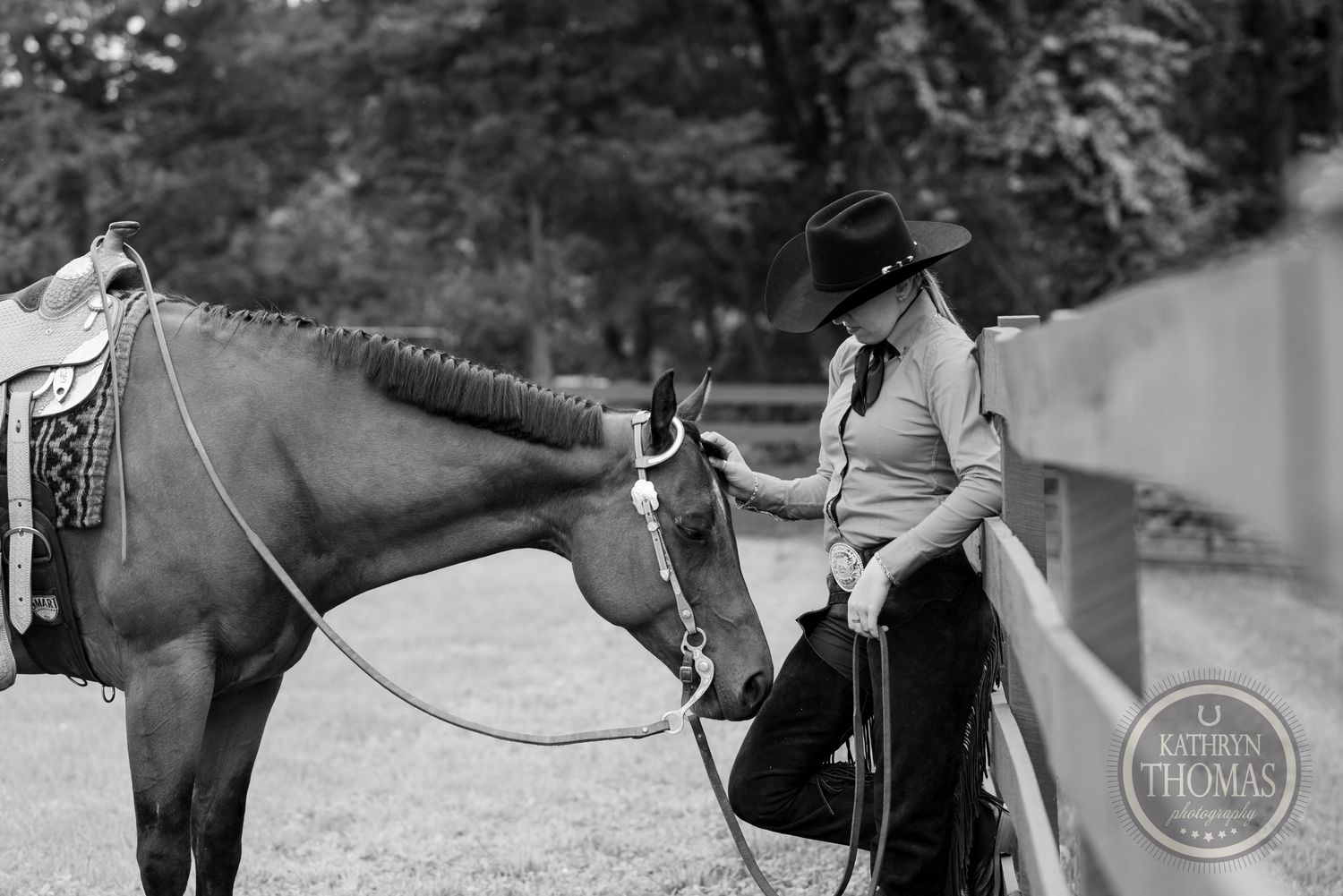 Black and white photo of horse and rider by fence in rustic setting.