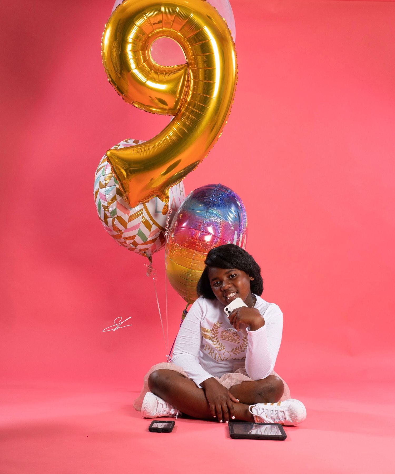Child poses playfully with large number 9 balloon and colorful decorations against pink background.
