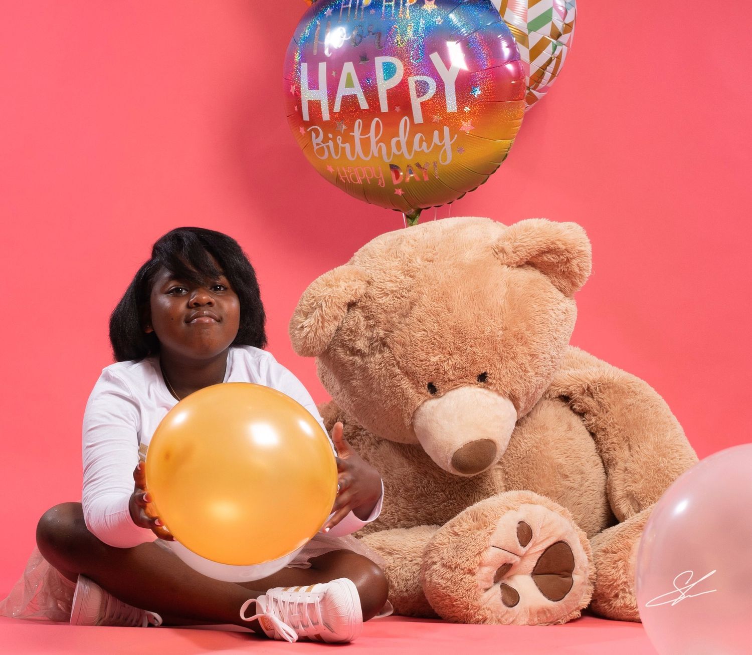 Child sits with yellow balloon and large teddy bear against pink background.