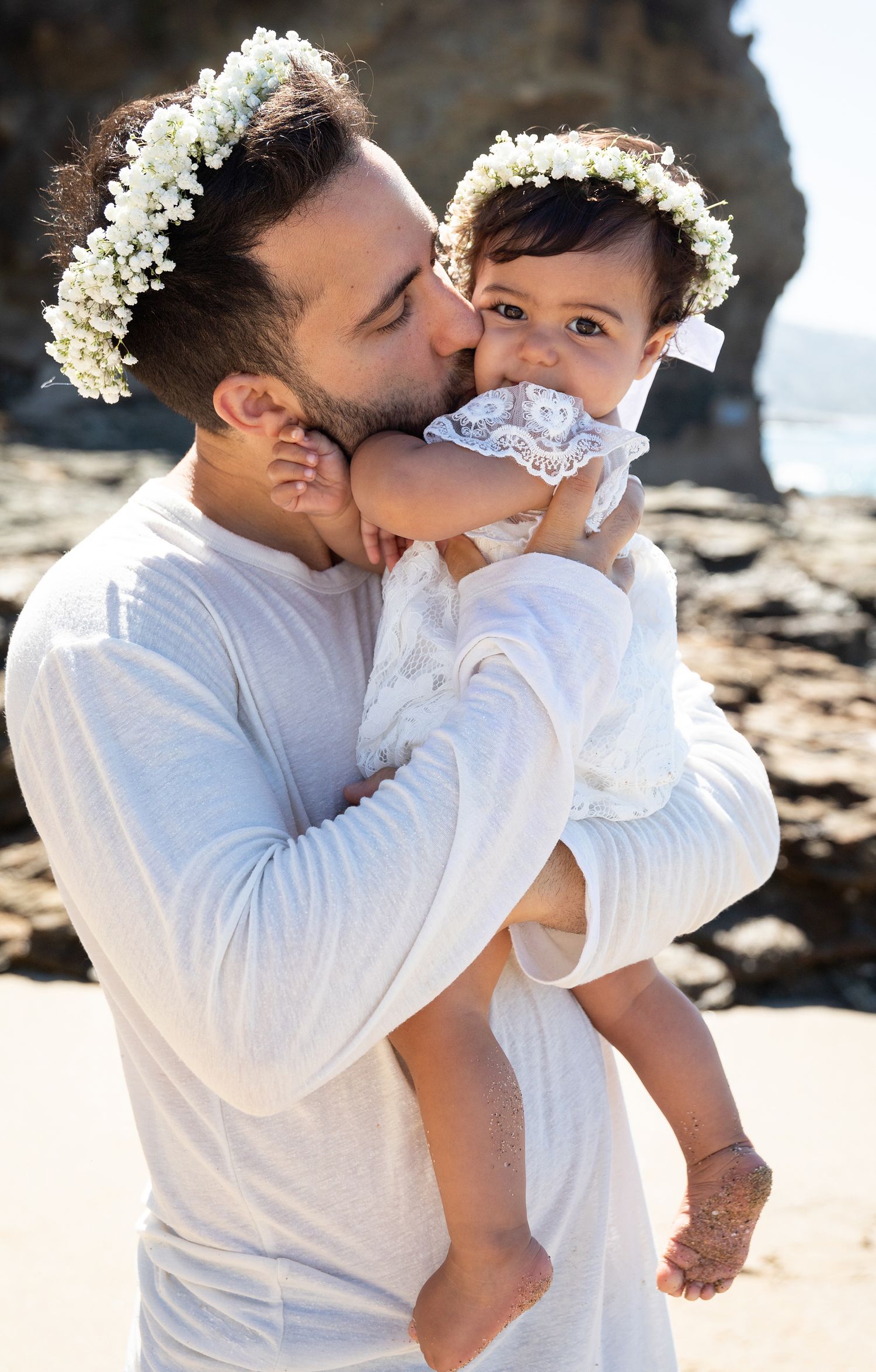 A tender moment between parent and baby wearing white outfits and floral crowns on a beach.