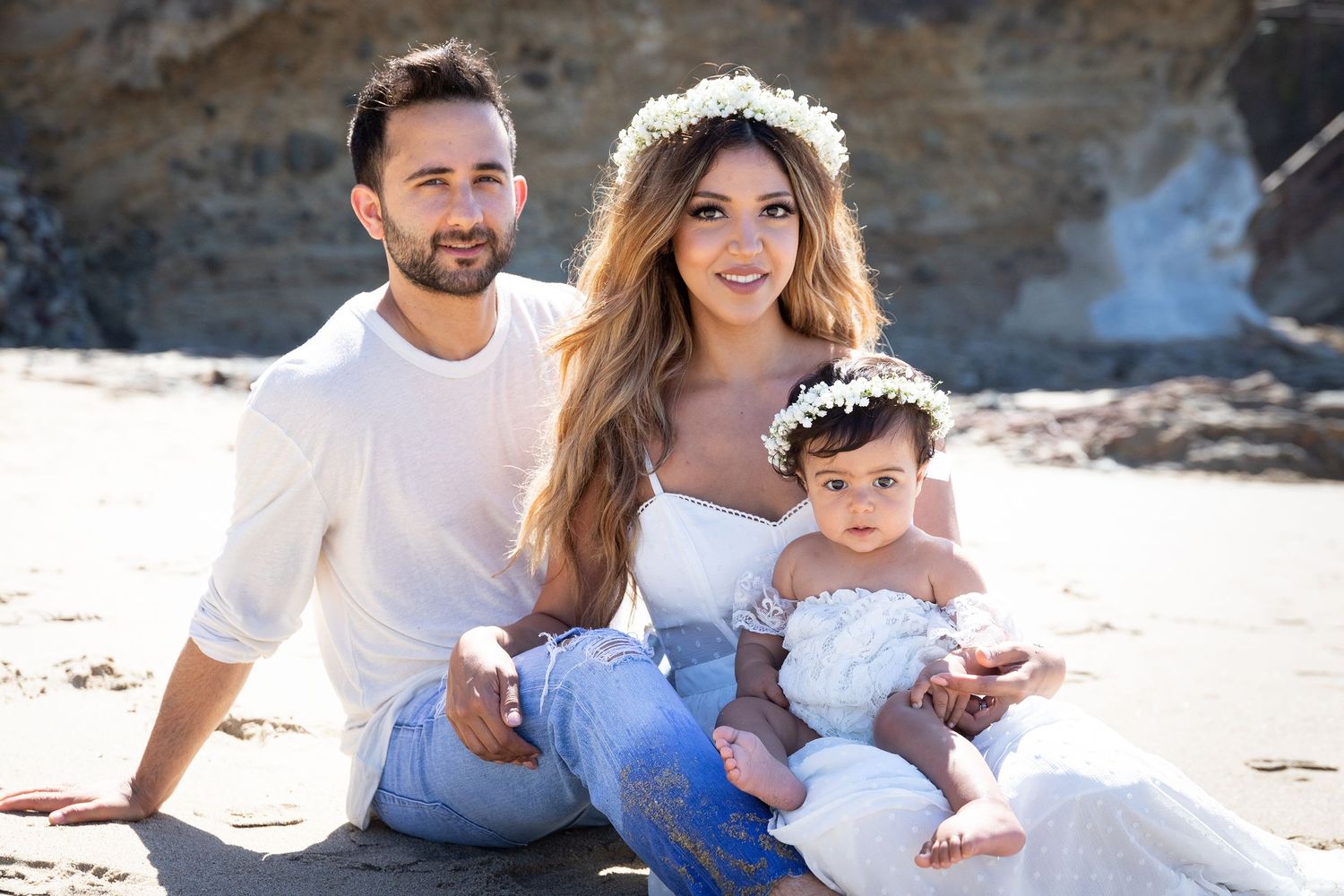 Family in white outfits sits together on sandy beach near rocky cliffs.