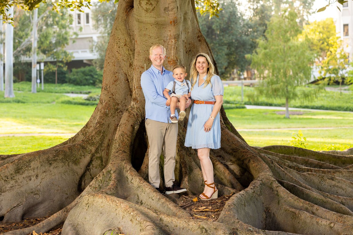 Family standing on large tree roots in a sunny park setting.