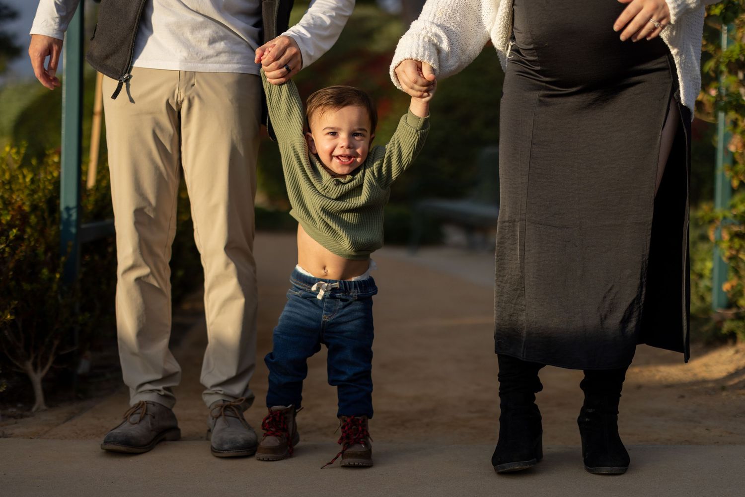 A toddler in a green sweater joyfully swings between two adults on a sunny afternoon.