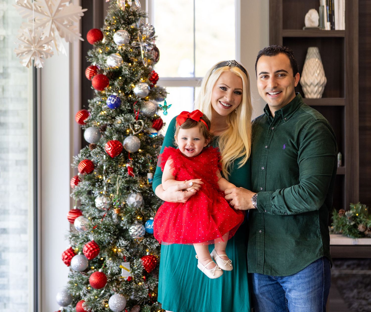 Family poses by decorated Christmas tree with baby in red dress.