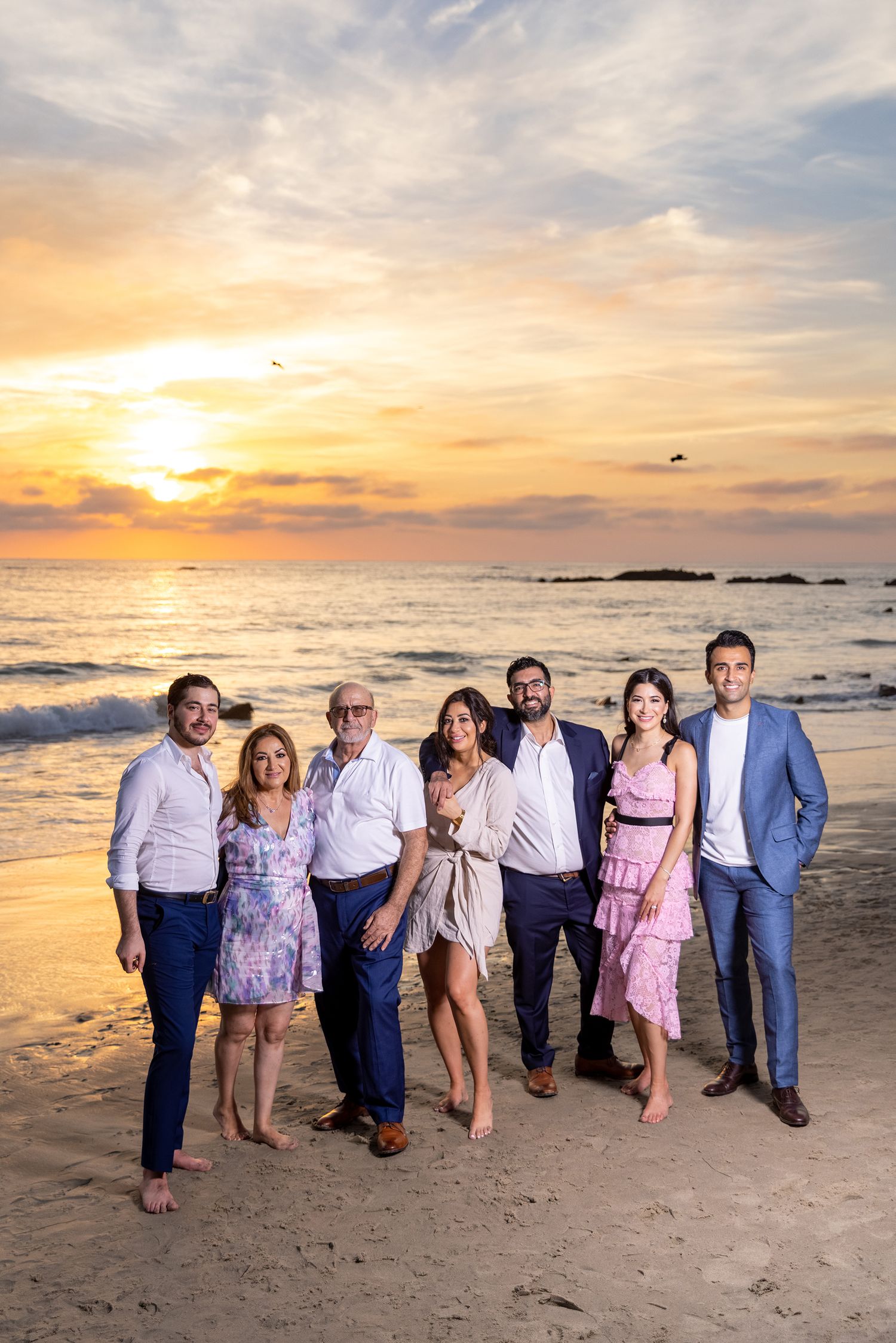 Group portrait of elegantly dressed people on beach at sunset with orange sky.