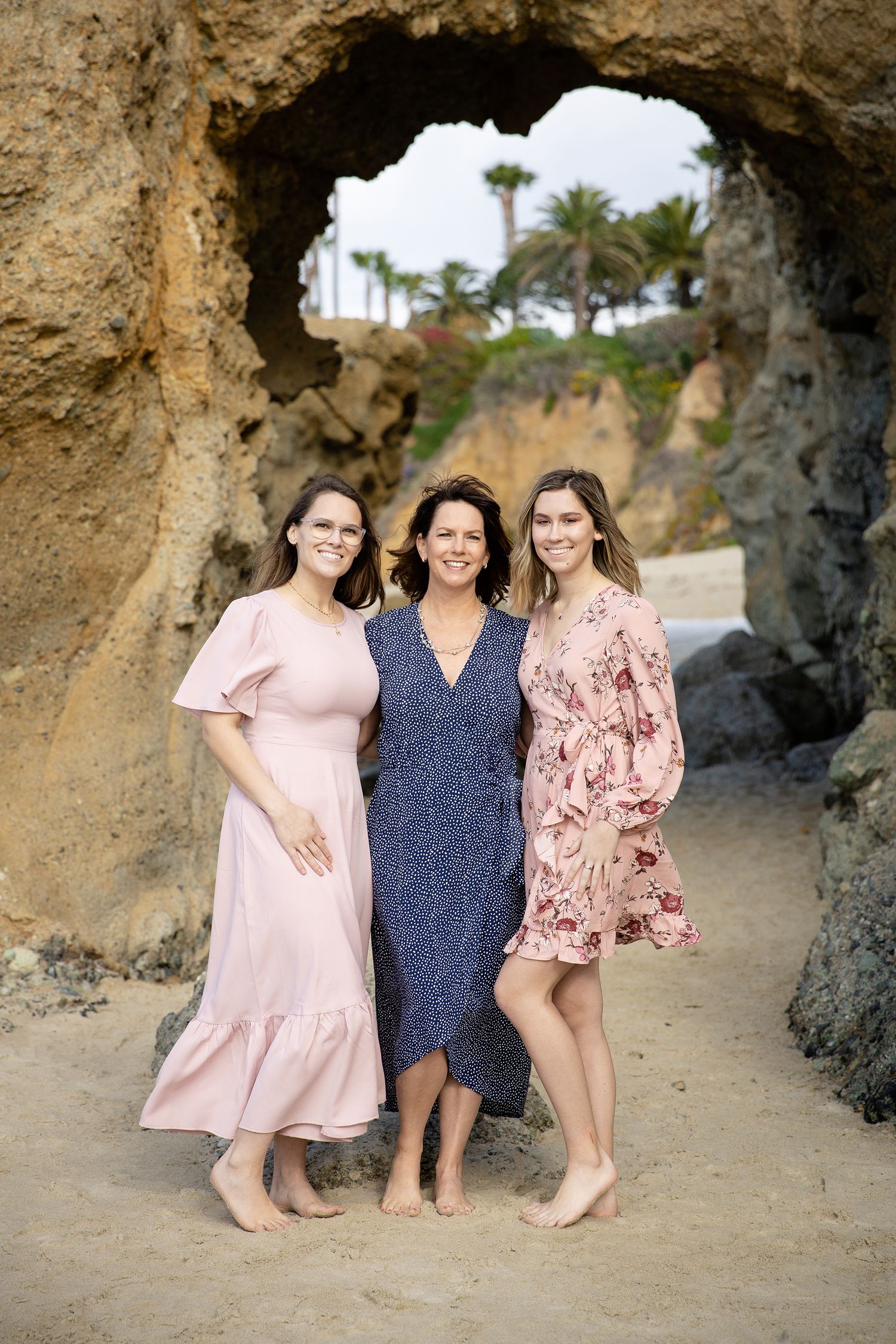 Three women in floral dresses pose inside natural rock arch formation at beach.