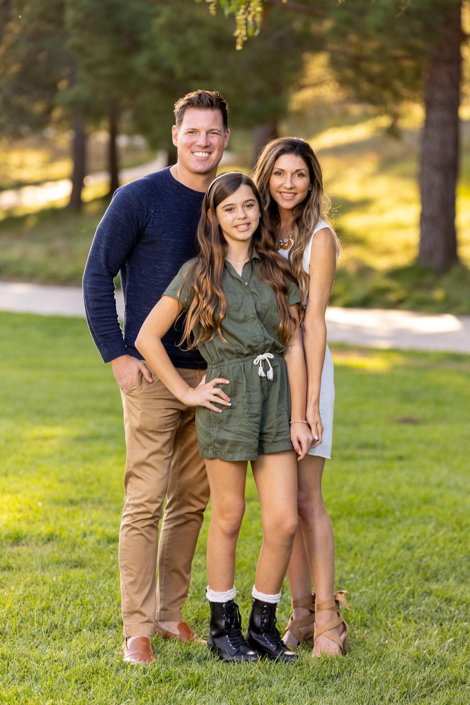 Family of three poses together on sunny grass hill in casual attire.