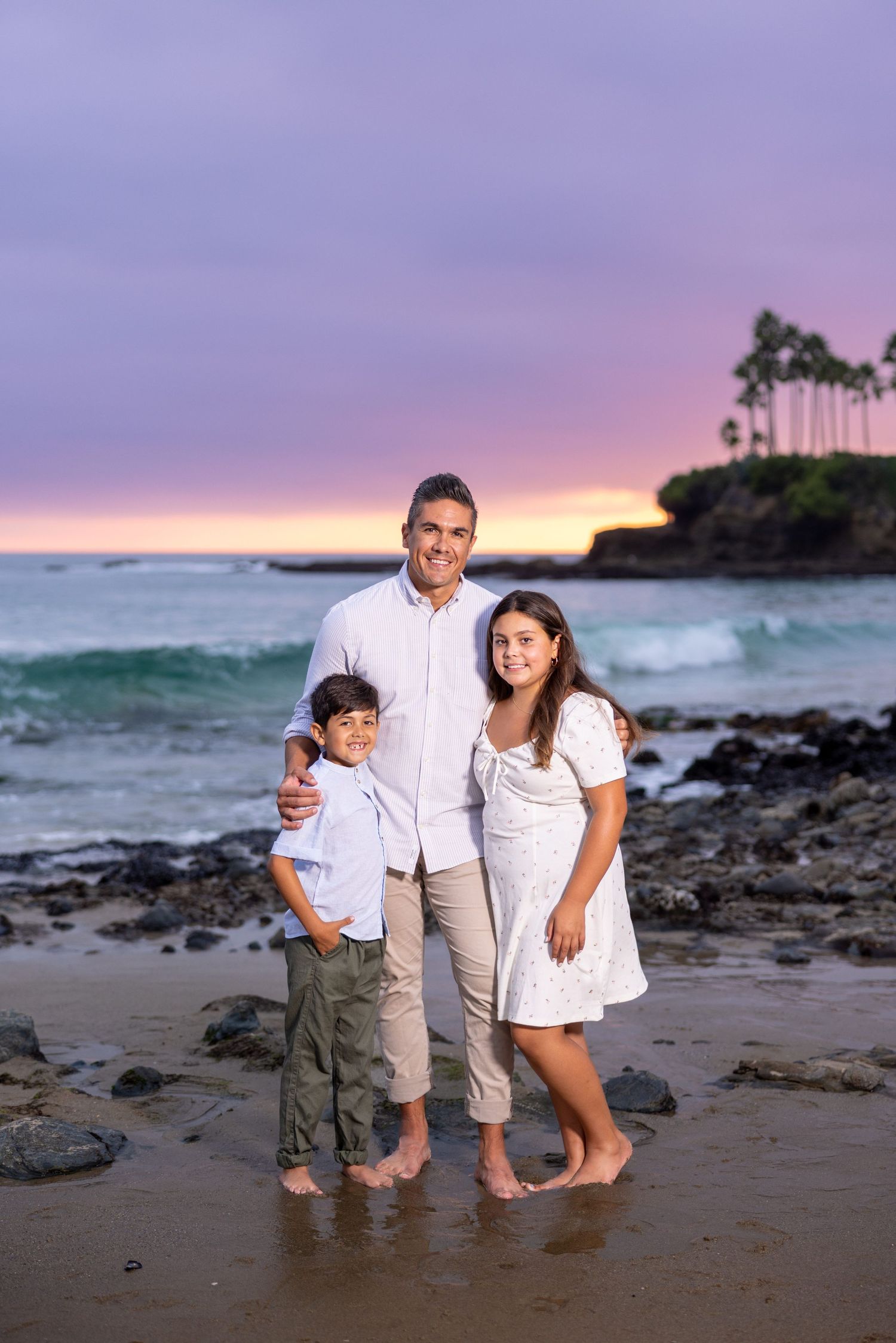 Three people pose together on a beach at sunset with waves and palm trees in the background.