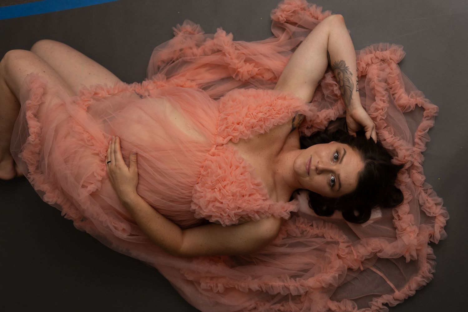Woman in flowing coral pink dress lying on dark floor in artistic pose.