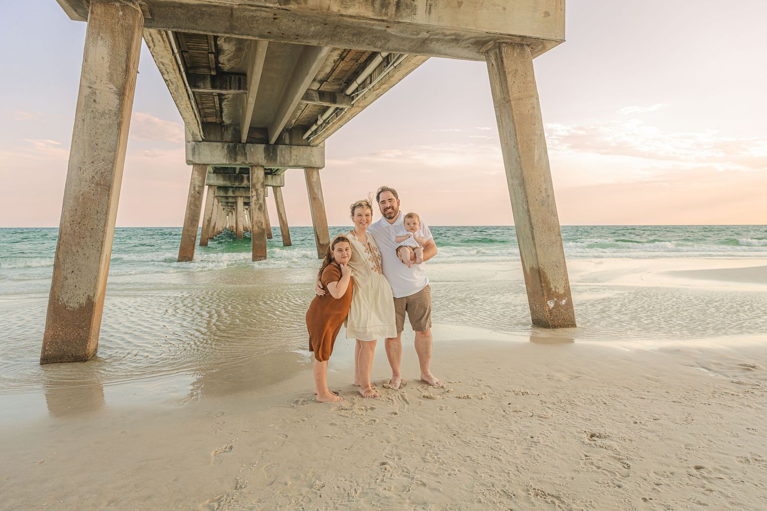 Family portrait session under a wooden pier at sunset with waves rolling in the background.