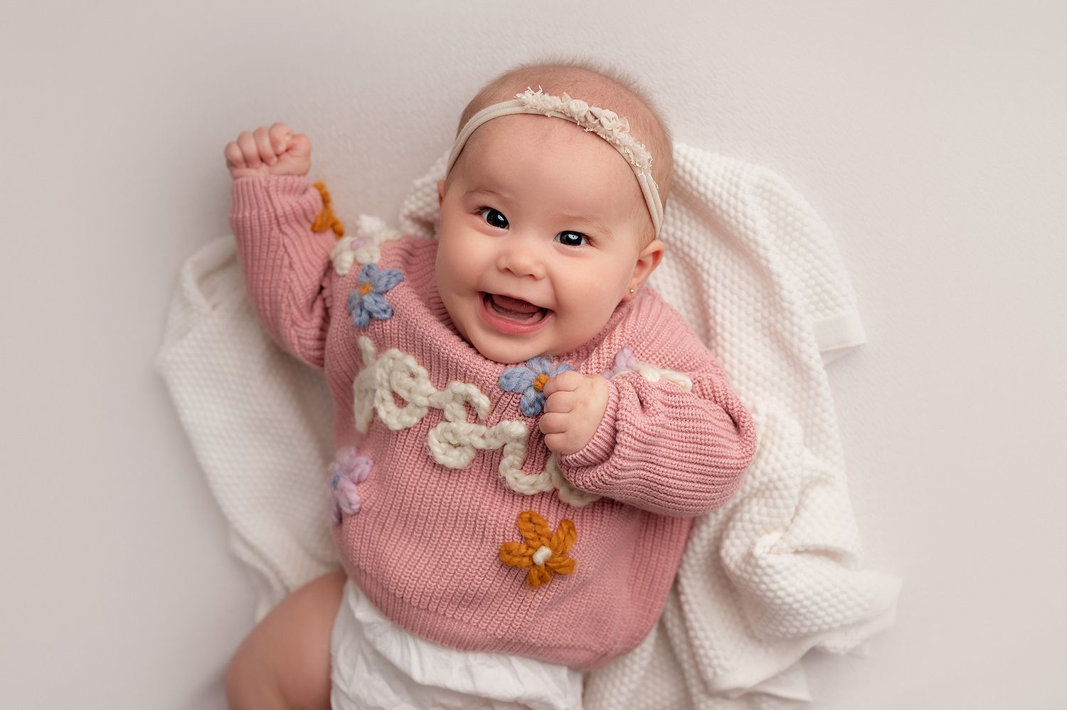 Baby wearing pink sweater and headband smiling while lying on white fabric.