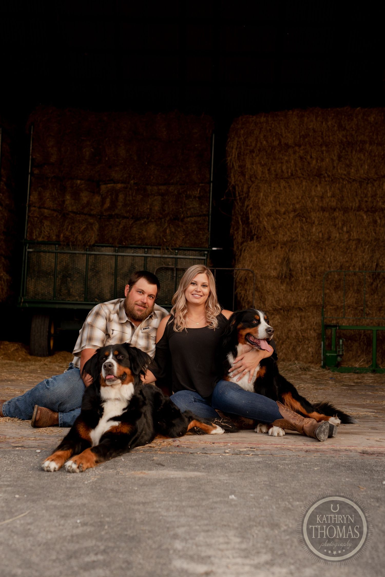 Couple sits with their Bernese Mountain Dogs in a rustic barn setting with hay bales in the background.