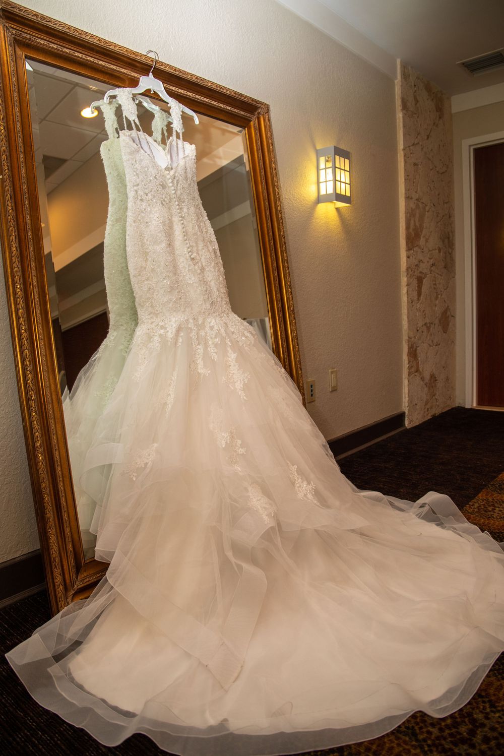 A beautiful white lace wedding dress with a flowing tulle skirt displayed on a hanger against gold-framed mirrors.