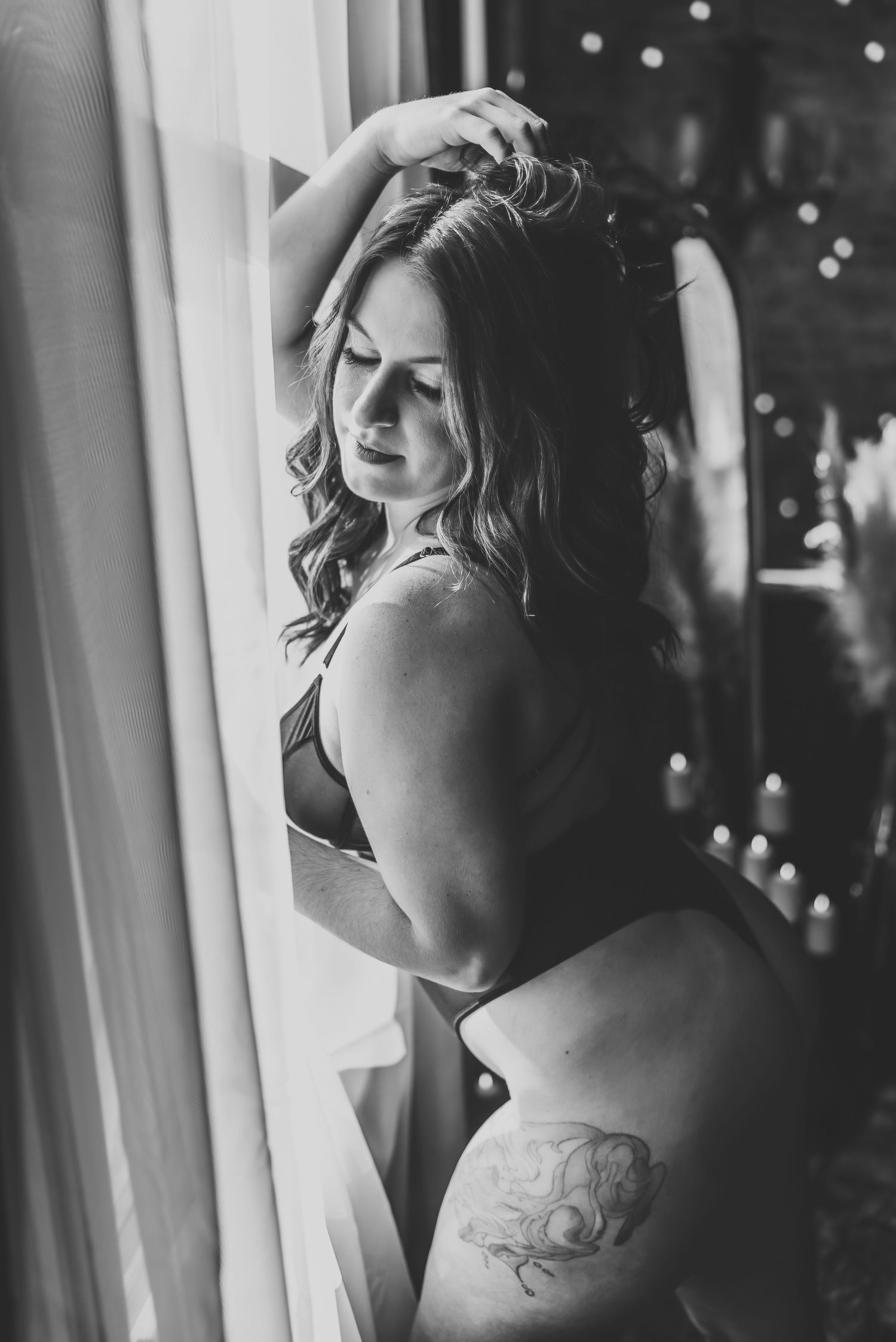 Black and white boudoir portrait by a window with twinkling lights in background.