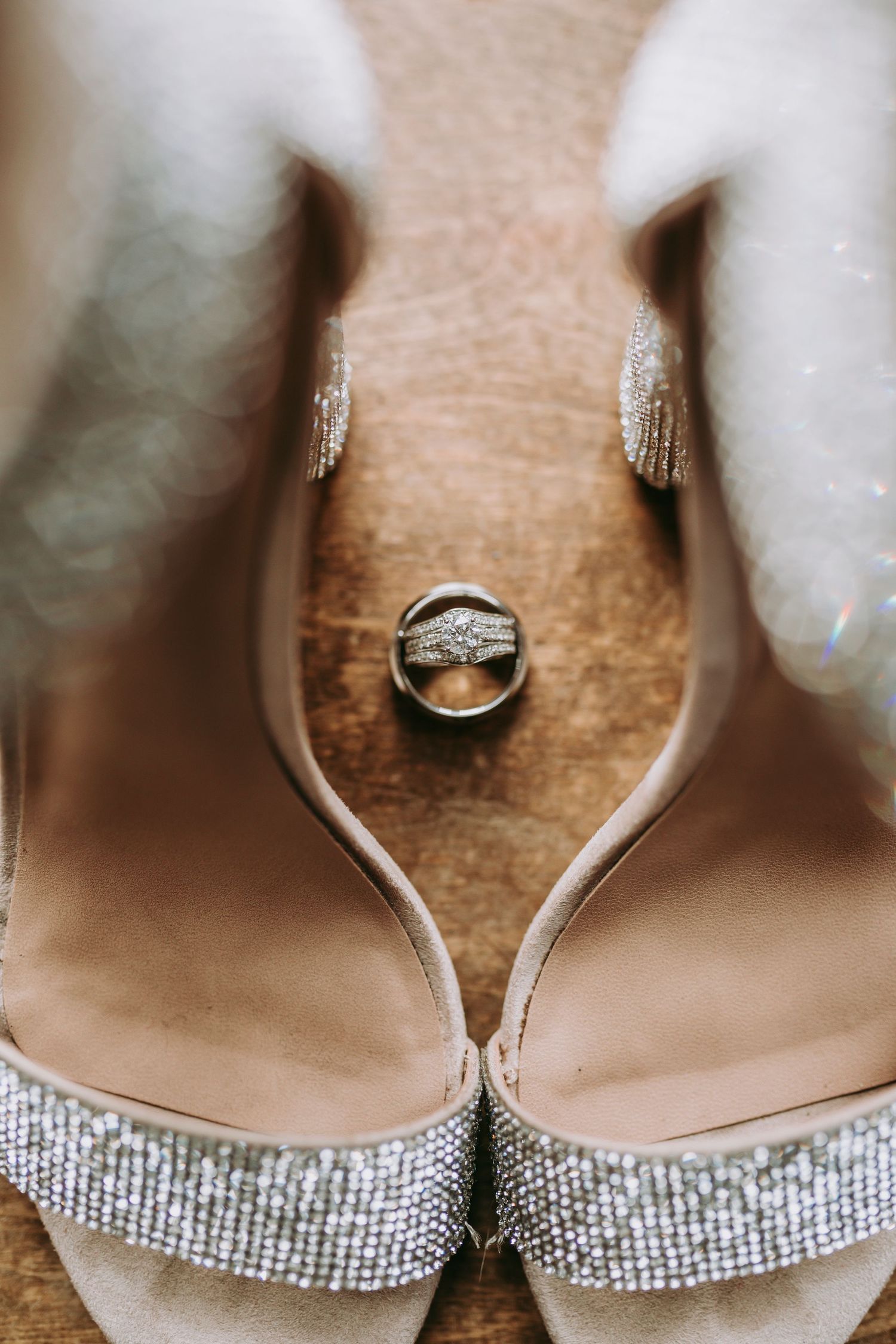 Sparkly wedding shoes with diamond ring balanced between them on wooden surface.