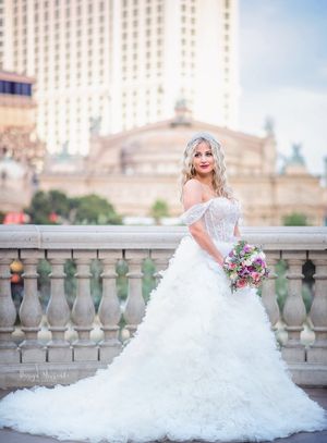 Bride in flowing white gown with bouquet poses on balcony with grand hotel architecture behind her.
