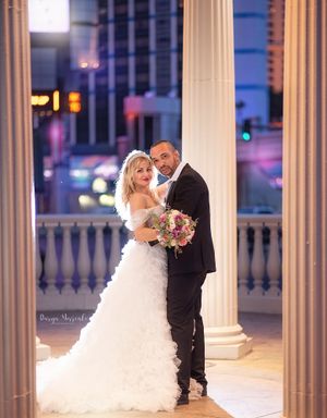 Wedding portrait between classic white columns at dusk with city lights twinkling in background.
