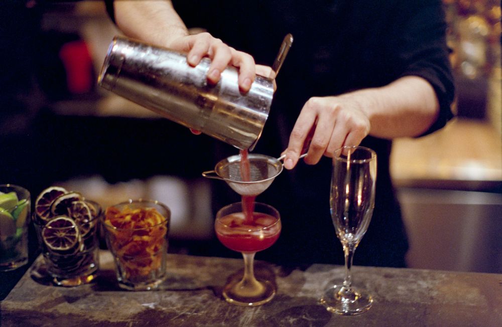 Hands pouring cocktails from a shaker into elegant glasses at a bar.
